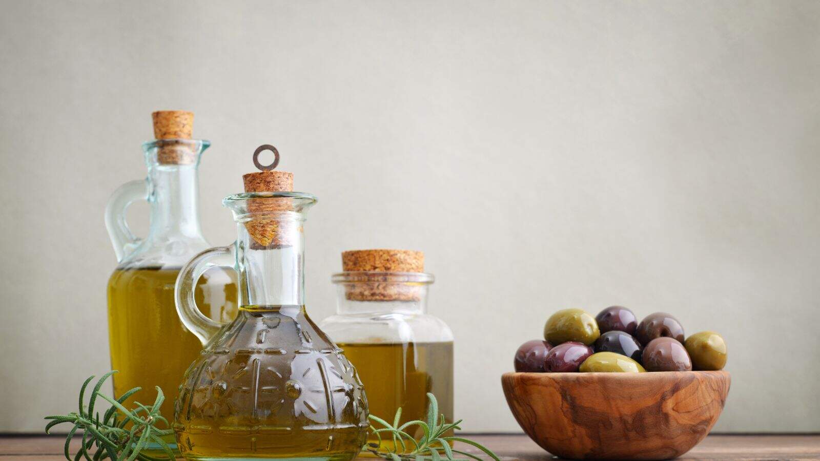 Three glass bottles filled with olive oil, each with a cork stopper, sit on a wooden surface next to a small wooden bowl containing green and black olives. Sprigs of rosemary are also present.
