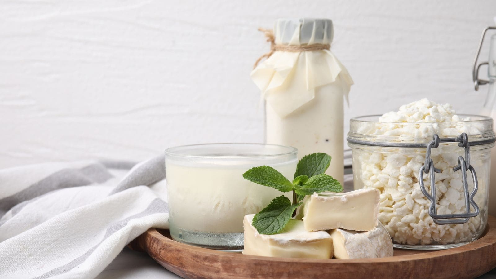 A wooden tray holds a glass of yogurt with a mint garnish, pieces of soft cheese, a jar of cottage cheese, and a bottle of milk with a fabric cover. A white cloth is placed beside the tray.