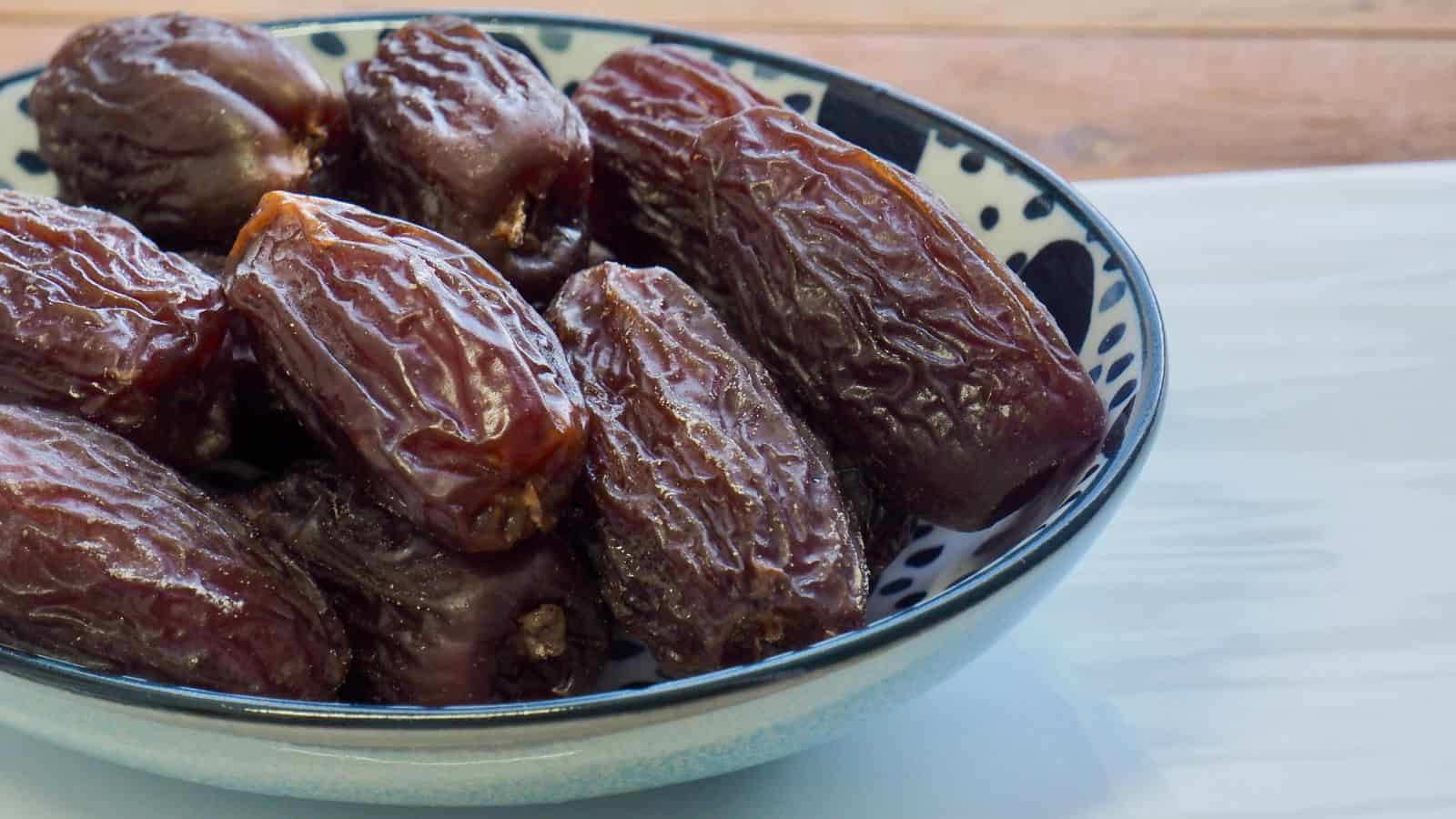 A blue and white patterned bowl filled with several whole dried dates sits on a white surface with a wooden background.