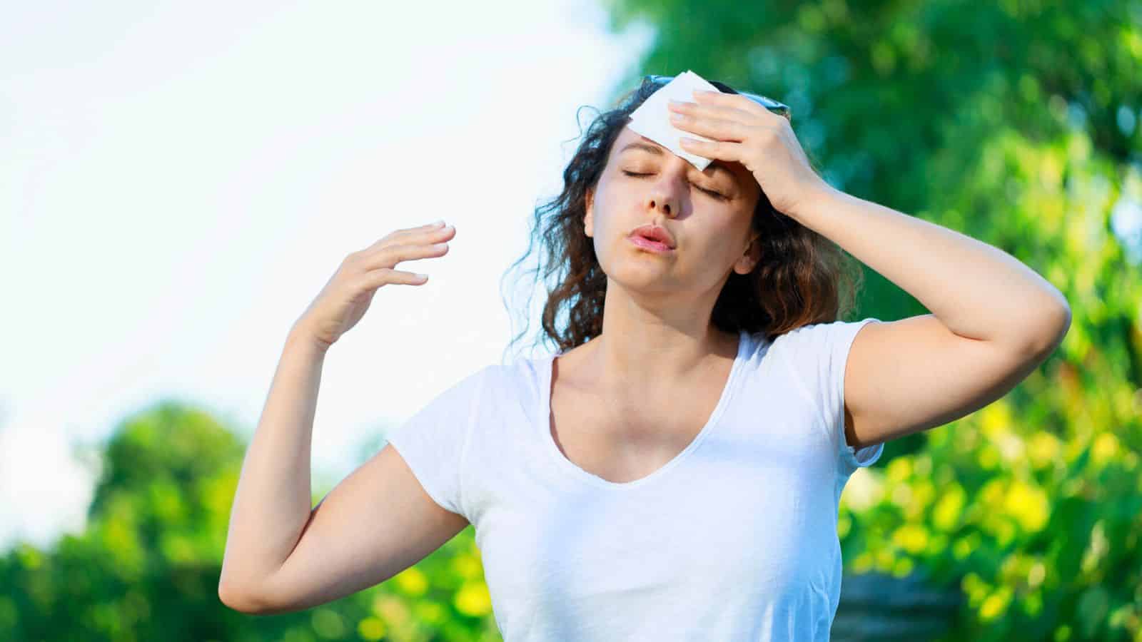 A person wearing a white shirt wipes their forehead with a cloth while standing outside, eyes closed and appearing overheated. Green foliage is visible in the background.