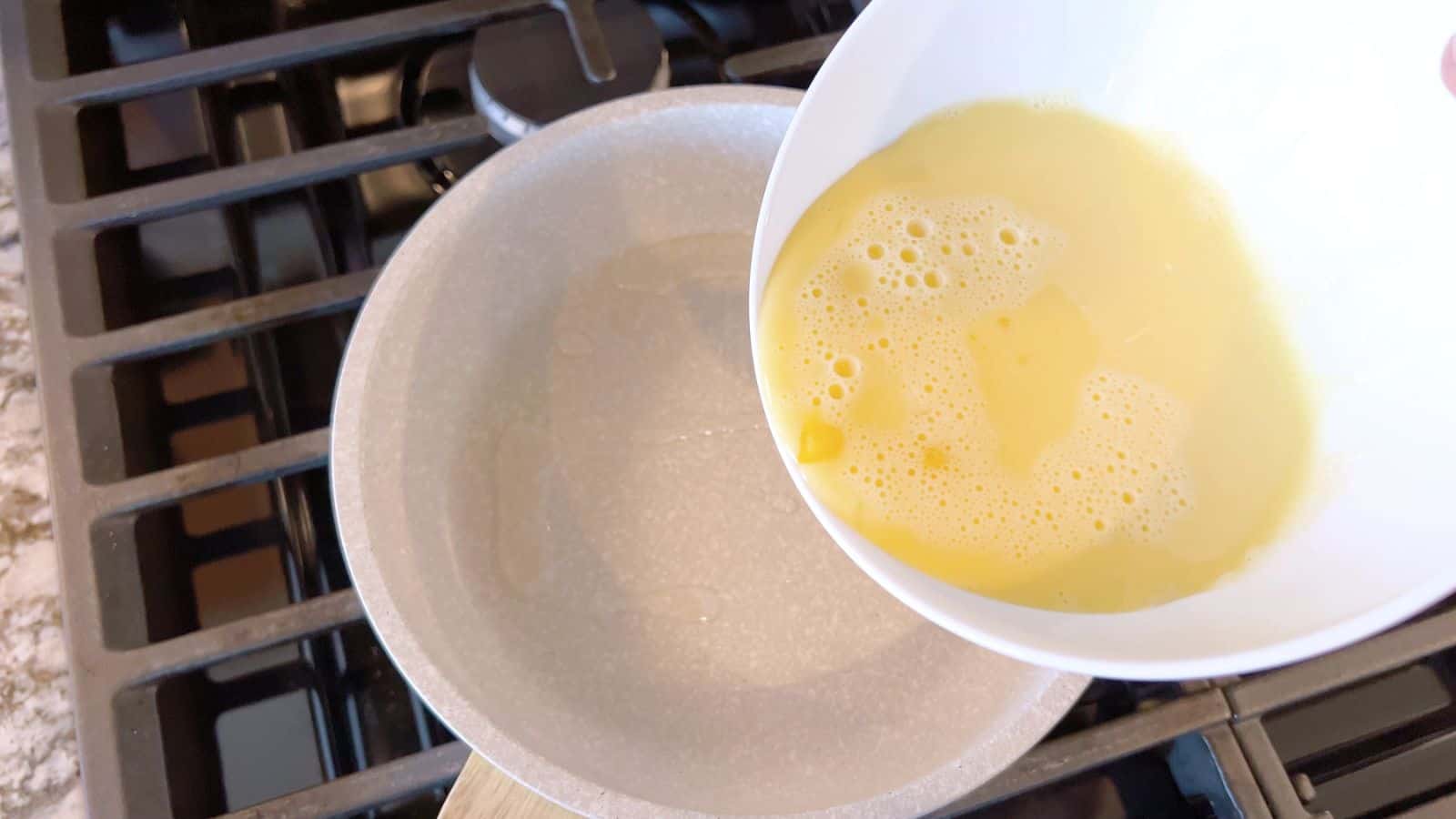 A hand pours beaten eggs from a white bowl into a light gray frying pan on a gas stove.