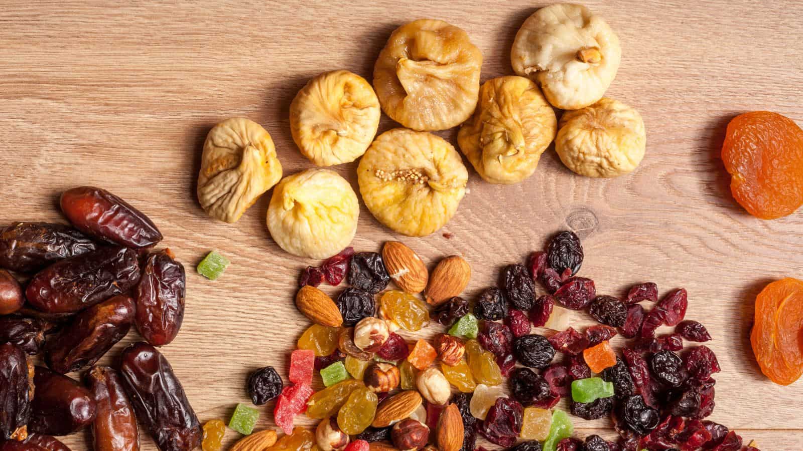 Assorted dried fruits and nuts, including figs, dates, apricots, raisins, almonds, and colorful diced dried fruit, arranged on a wooden surface.
