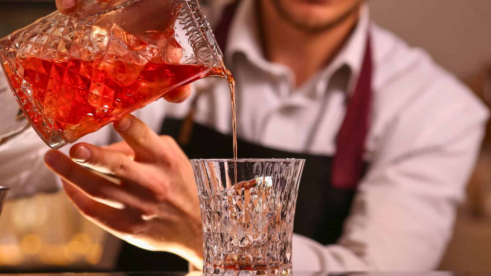 A person wearing an apron pours a red cocktail from a glass mixing pitcher with ice into a crystal glass, both held in their hands.