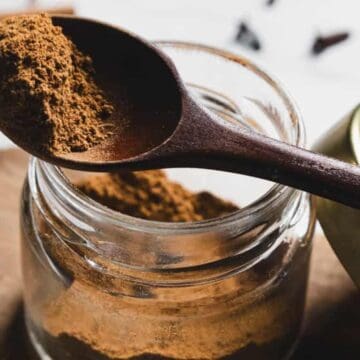 A wooden spoon holds ground cinnamon over an open glass jar filled with more cinnamon. The jar's metal lid rests on the table beside it, and a cinnamon stick is blurred in the background.