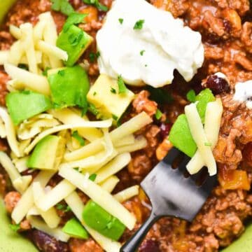 A bowl of chili with ground meat, beans, and tomato sauce, topped with shredded cheese, diced avocado, and a dollop of sour cream. A fork rests inside the bowl. The bowl is on a green napkin.