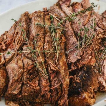 A white oval plate holds slices of cooked beef roast garnished with several sprigs of fresh thyme. The meat appears tender with visible seasoning and herbs on top.