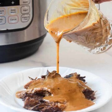 A hand pours brown gravy from a glass container over shredded beef on a white plate. An electric pressure cooker sits in the background on a white surface.