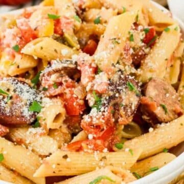 A white bowl filled with penne pasta, sliced sausage, diced tomatoes, and sprinkled grated cheese, garnished with chopped parsley. A fork and a garlic bulb are visible nearby.