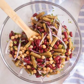 A glass bowl filled with mixed bean salad, including kidney beans, garbanzo beans, green beans, and sliced red onions, with a wooden spoon resting inside. A striped cloth is partially visible on the right.