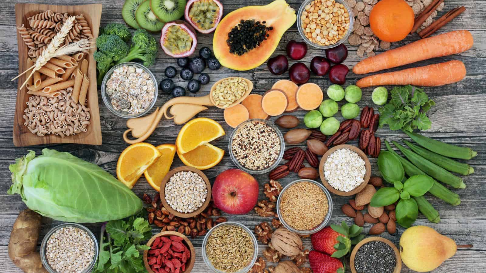 An assortment of fresh fruits, vegetables, grains, legumes, nuts, and seeds arranged on a wooden surface, including pasta, berries, oranges, carrots, kiwi, papaya, apples, leafy greens, and various seeds in bowls.