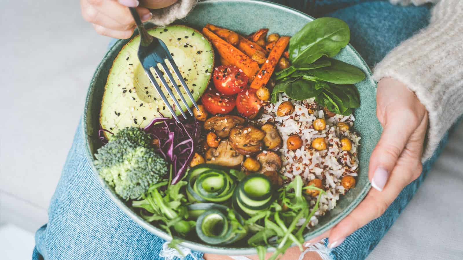 A person holds a bowl containing broccoli, sliced avocado, carrots, cherry tomatoes, spinach, rice, chickpeas, sautéed mushrooms, cucumber ribbons, and leafy greens, with a fork in hand resting on their knee.