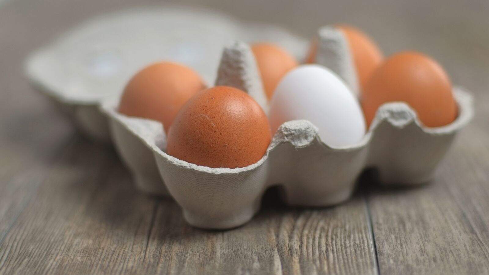 A close-up of a cardboard egg carton holding five eggs, four brown and one white, placed on a wooden surface. The focus is on the eggs in the foreground.