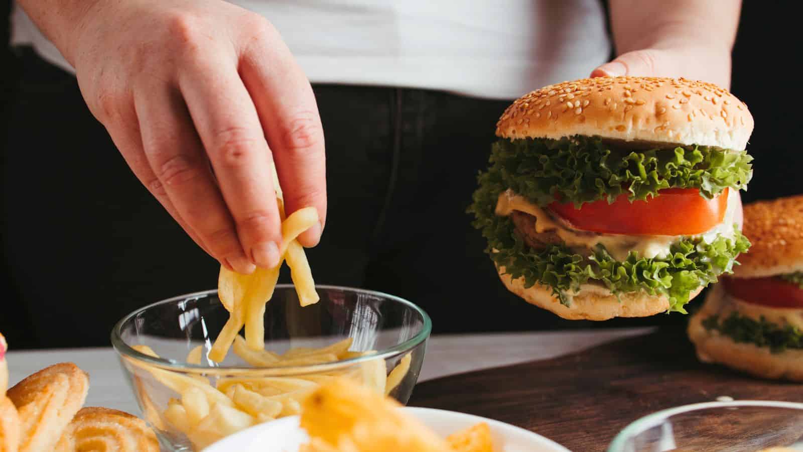 A person holds a large hamburger with lettuce, tomato, and sauce in one hand while picking up French fries from a glass bowl with the other hand. Additional food items are visible on the table.