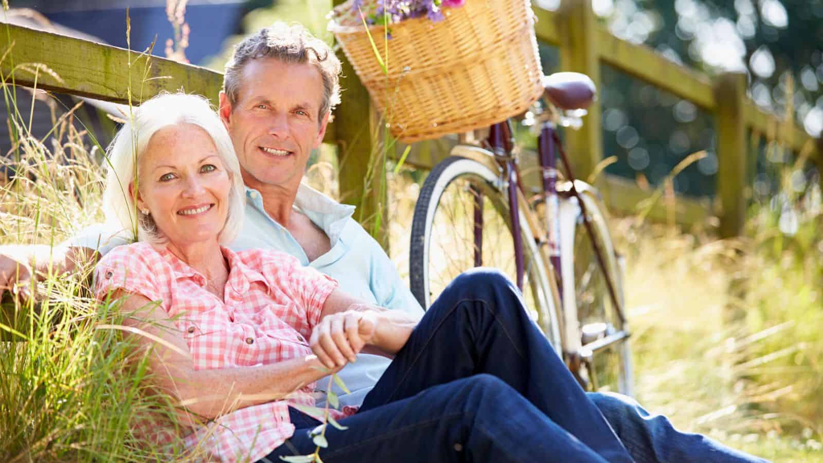 An older couple sits on grass, leaning against a wooden fence next to a bicycle with a basket of flowers. They are smiling and dressed casually, with sunlit greenery in the background.