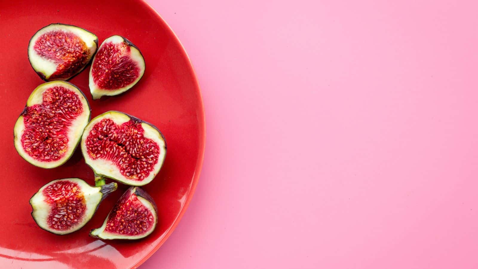A red plate with six halved figs is placed on a pink surface. The figs have a dark purple skin and red, seeded interiors. The right side of the image has empty pink space.