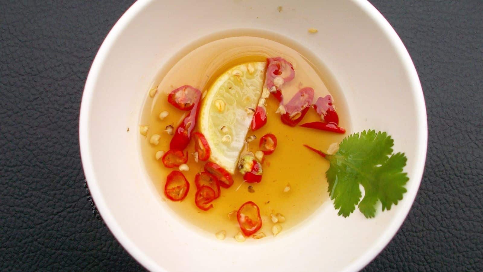 A white bowl containing light brown sauce with red chili slices, a lime wedge, and a sprig of cilantro on a dark textured surface.