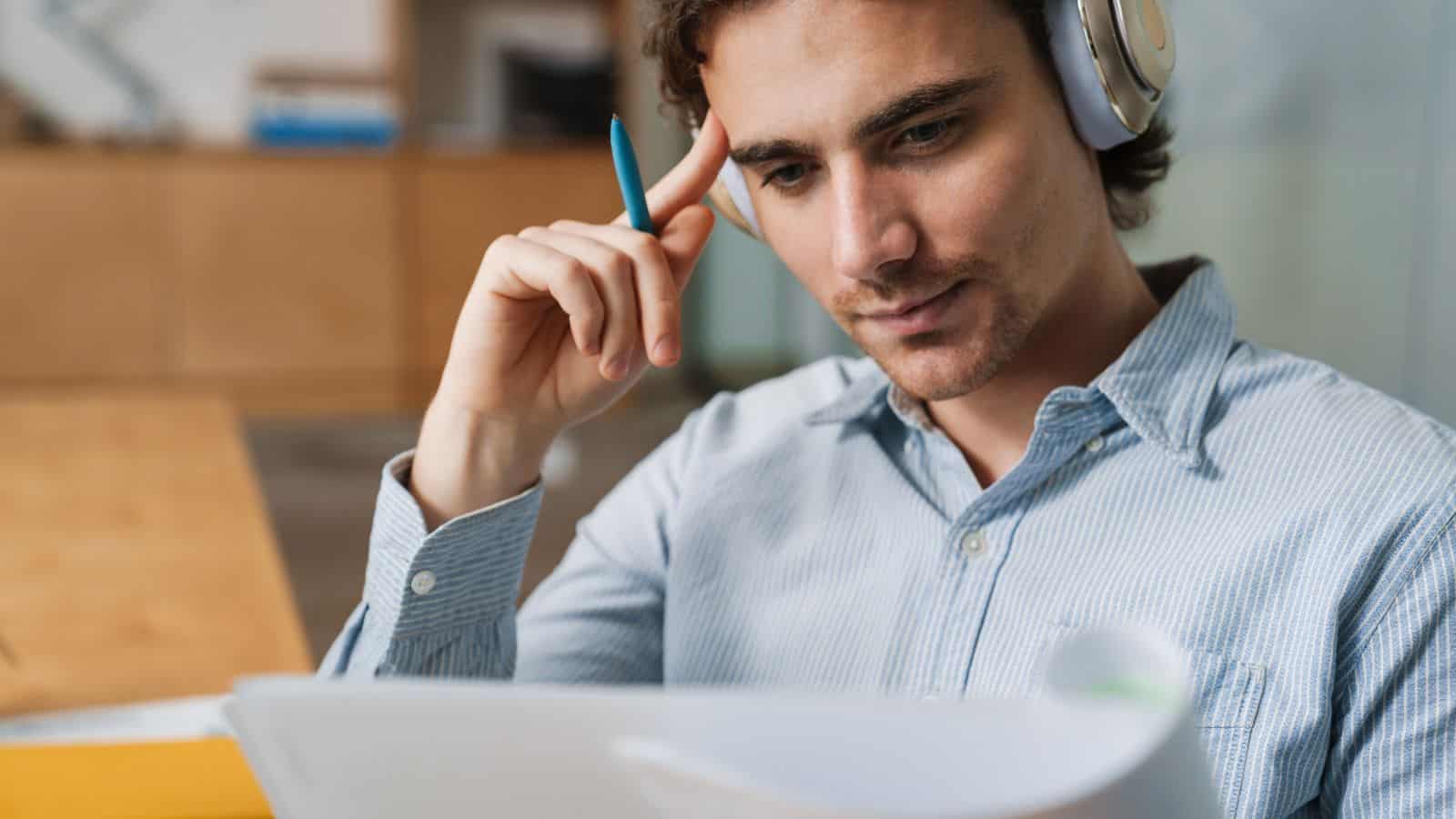 A person wearing headphones and a light blue striped shirt holds a pen and looks intently at a document or notebook, seated at a desk in a well-lit room with shelves in the background.