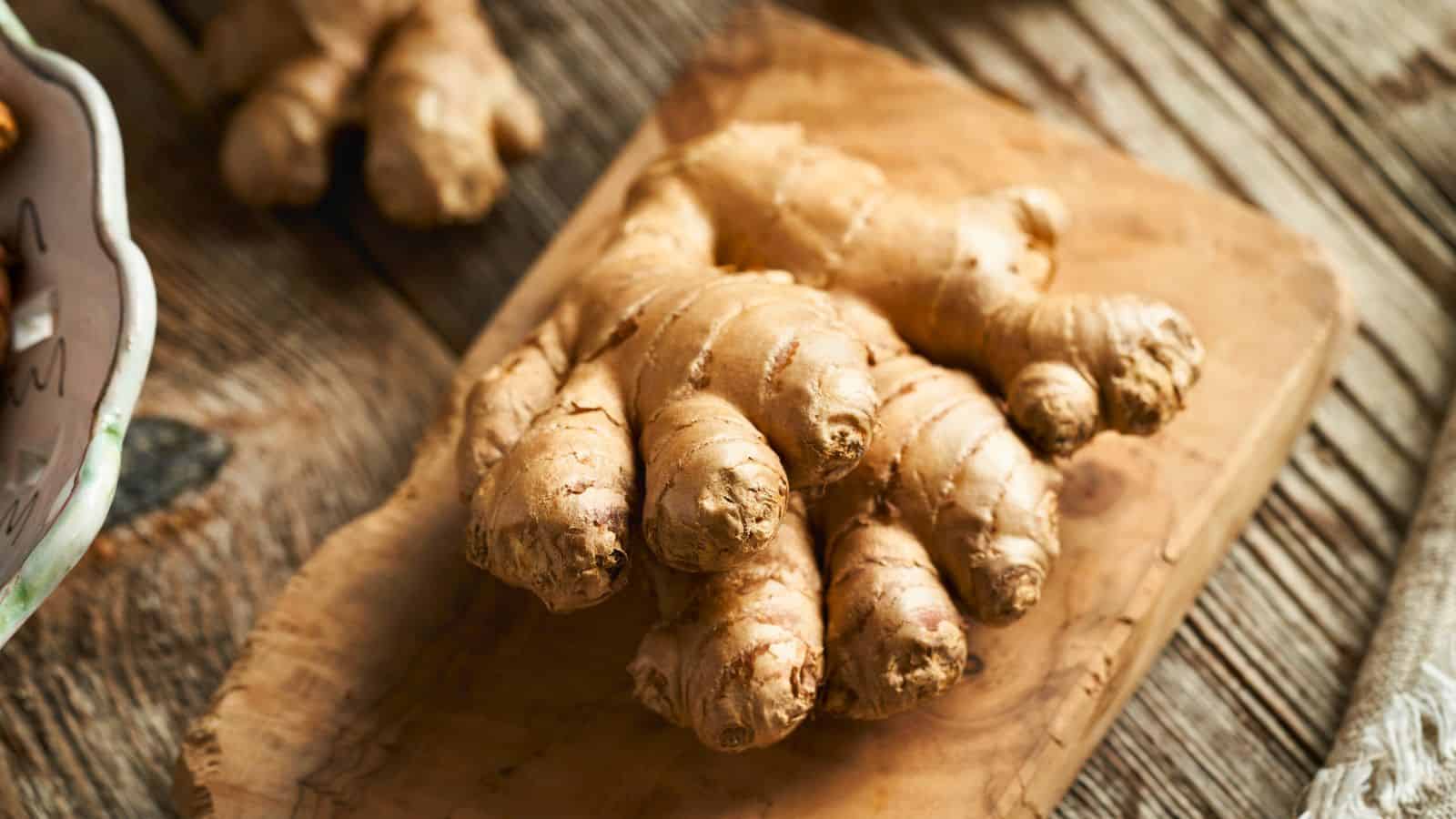 A fresh ginger root with light brown skin sits on a wooden cutting board. The background shows parts of another ginger root and a rustic wooden surface.