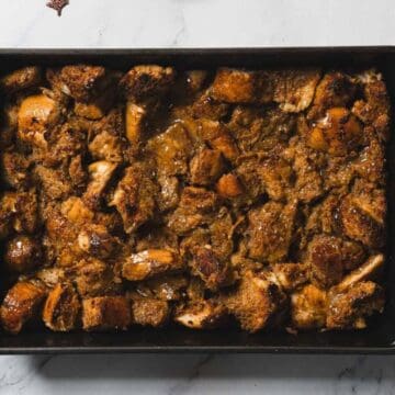 A rectangular baking dish filled with baked bread pudding sits on a marble surface, surrounded by autumn-themed decor such as a pine cone, cinnamon sticks, and a brown cloth.