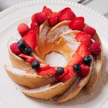 A Bundt cake topped with sliced strawberries, whole blueberries, and a dusting of powdered sugar sits on a white plate. A bowl of blueberries and a decorative plate are visible in the background.