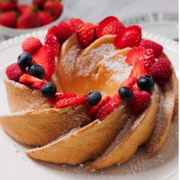 A Gluten-Free Angel Food Cake dusted with powdered sugar is topped with fresh strawberries, raspberries, and blueberries, displayed on a white plate with a bowl of strawberries in the background.