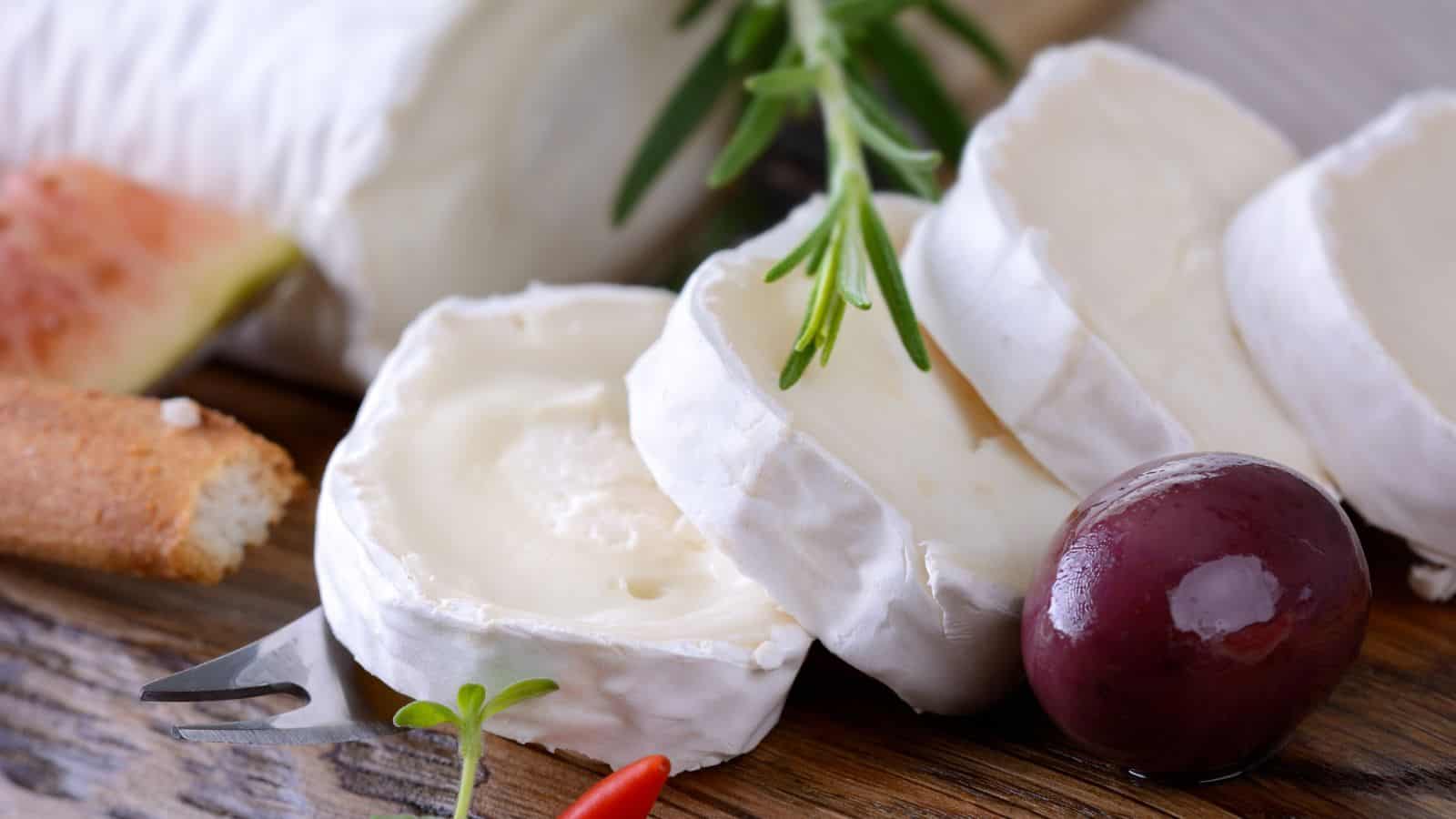 Slices of soft white cheese, possibly brie or goat cheese, rest on a wooden board with a sprig of rosemary, a dark olive, part of a baguette, and a piece of fig in the background.