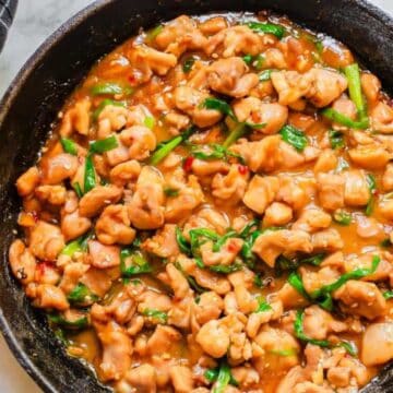 A cast iron skillet filled with cooked pieces of chicken and green onions in a brown sauce, with a black and white checkered cloth on the side.