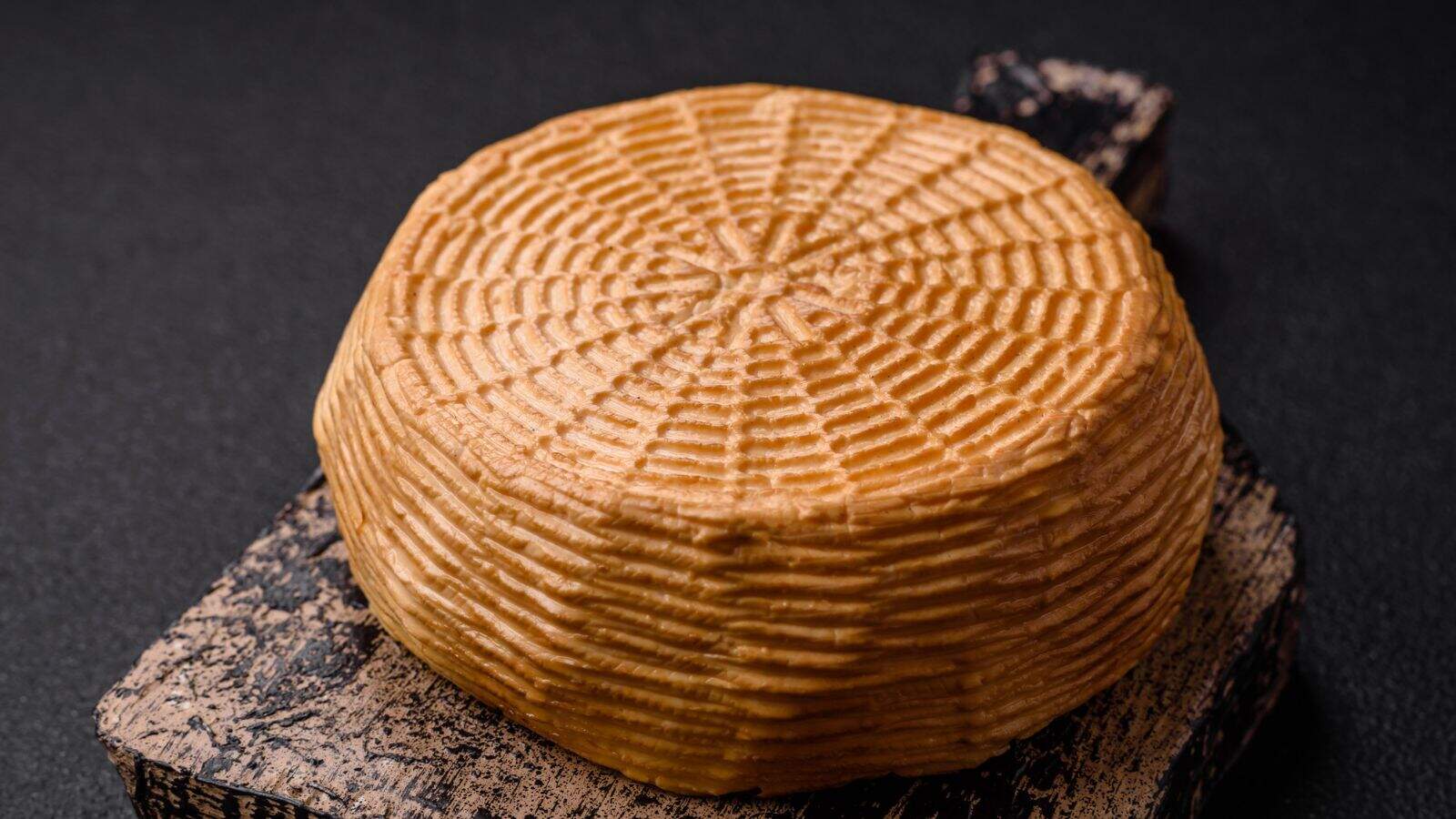 A round wheel of cheese with a textured, patterned rind sits on a rustic wooden board against a dark background.