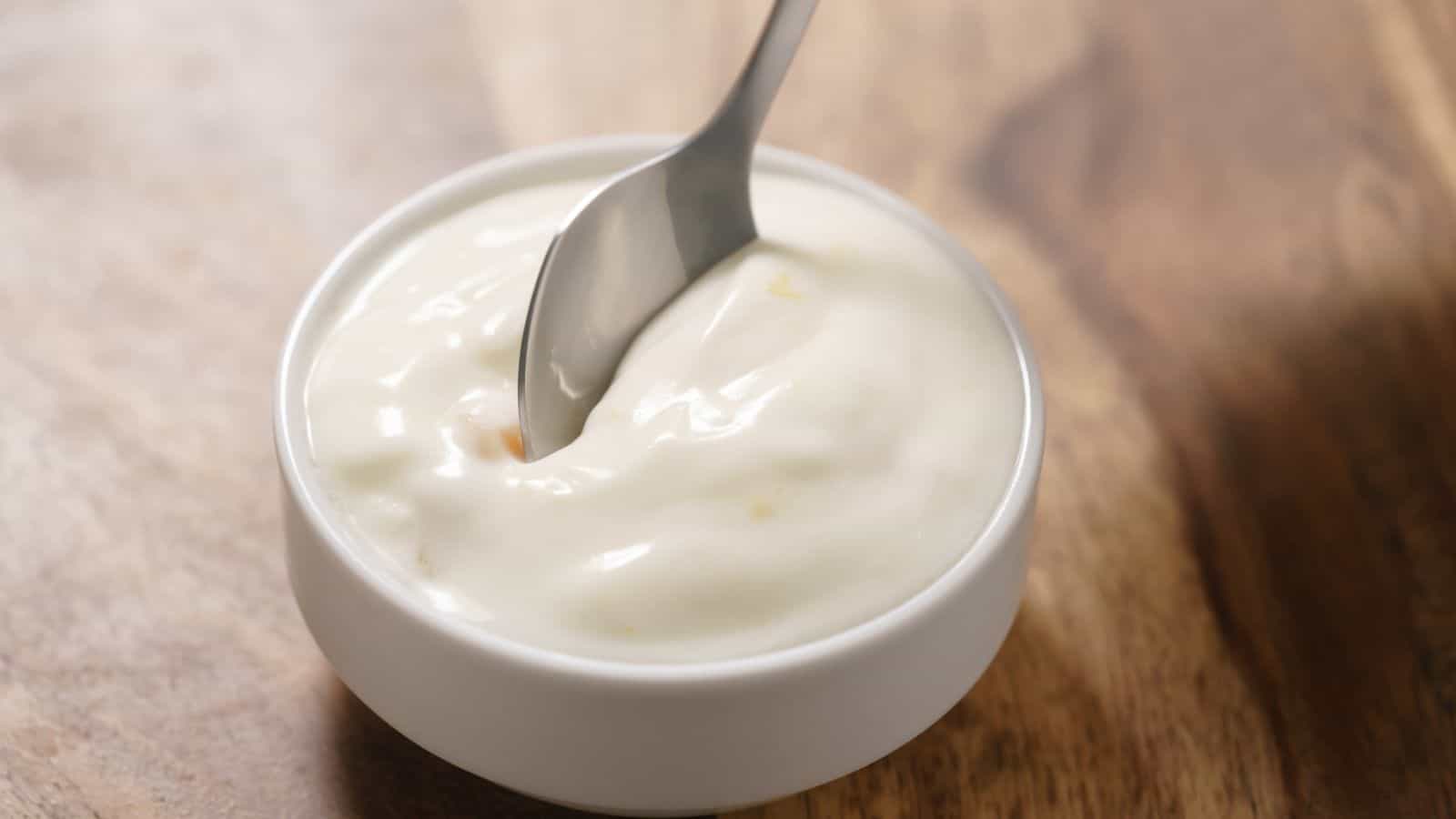 A metal spoon is dipped into a small white bowl filled with thick, creamy white yogurt, set on a wooden surface.