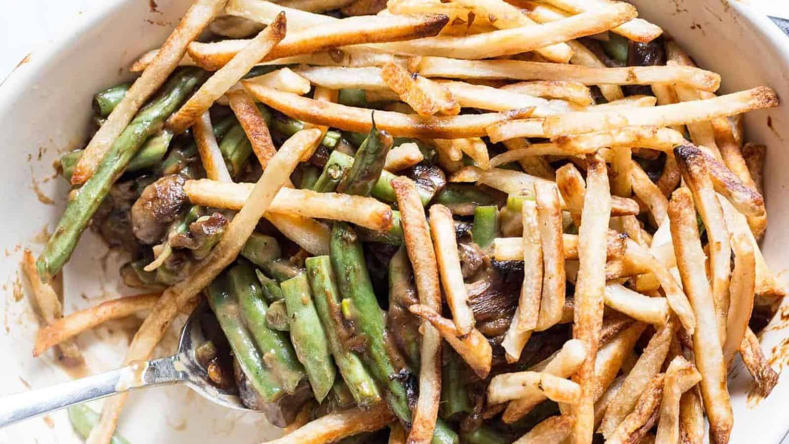 A casserole dish filled with cooked green beans and thin, crispy French fries, with a spoon lifting a portion of the mixture. Some fries and beans are coated in a dark sauce.