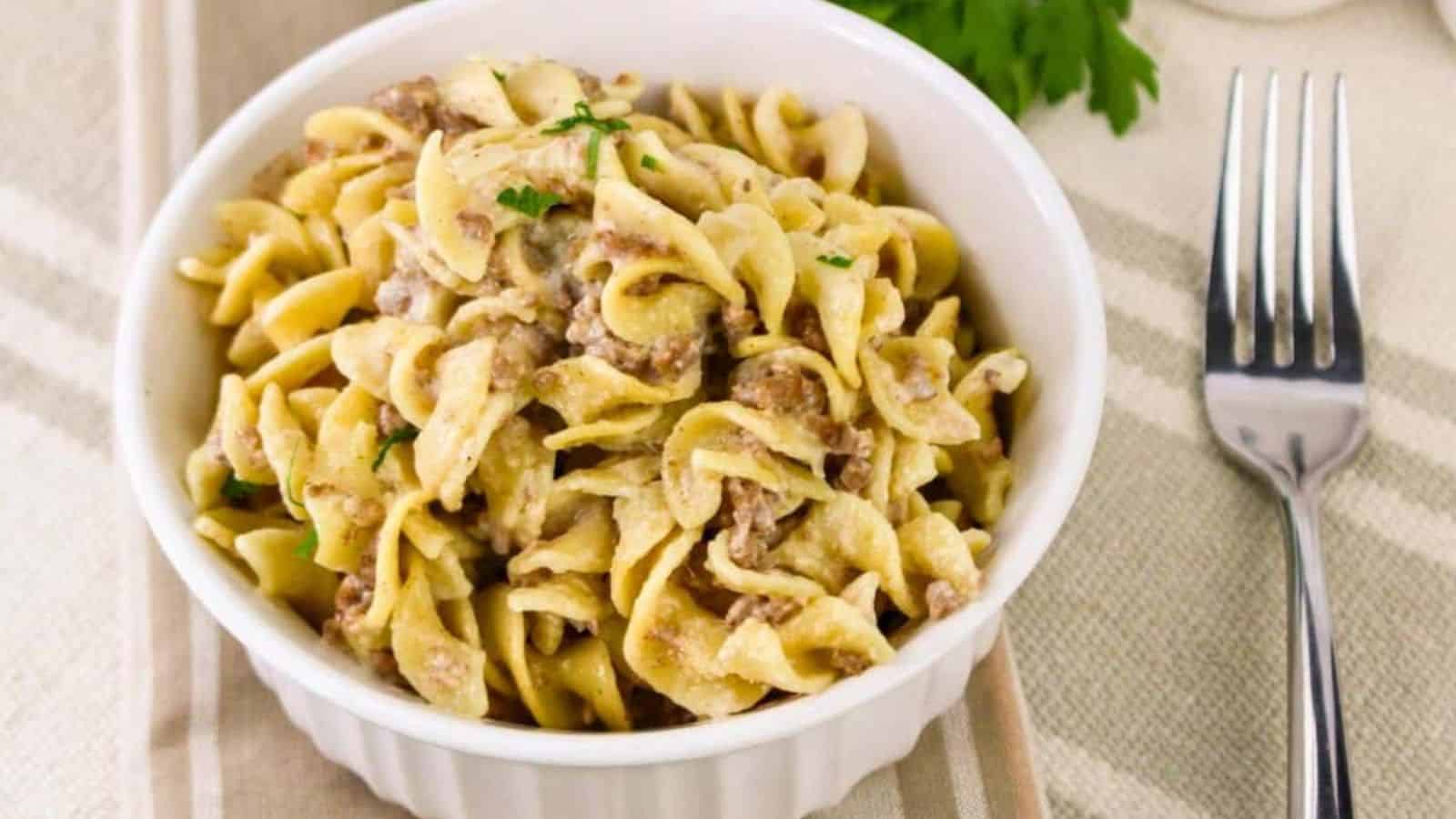 A bowl of creamy beef stroganoff with egg noodles on a striped cloth next to a fork and a sprig of parsley.