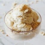 A glass dessert bowl filled with several scoops of halva tahini ice cream, topped with a light sprinkling of crumbled cookies on a white surface. Some crumbs are scattered around the bowl.