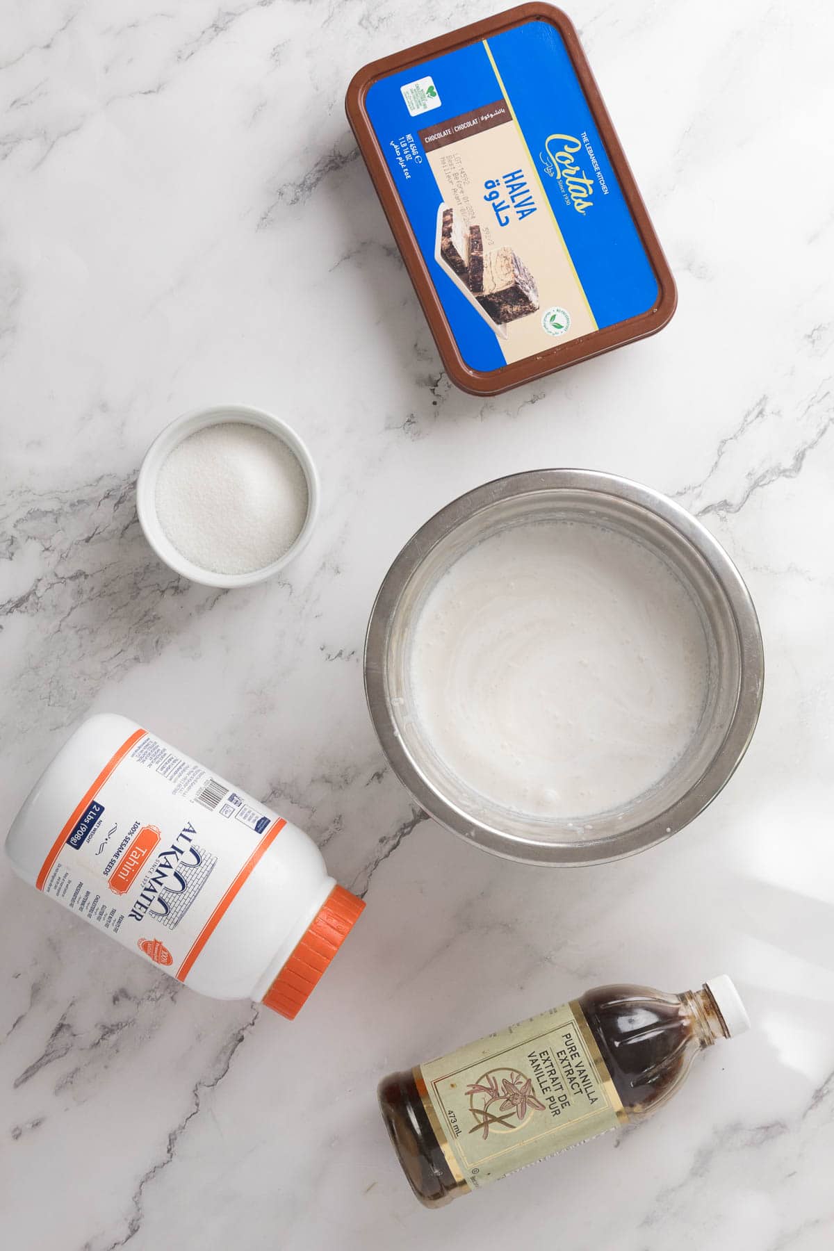 A bowl of white ice cream mixture, a container of Halva Tahini Ice Cream, a small bowl of sugar, vanilla extract, and xanthan gum are arranged on a white marble surface.