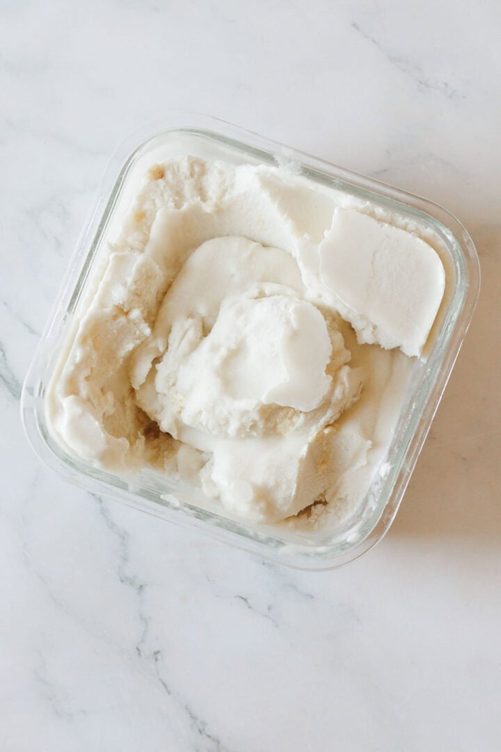 A square glass container filled with white, creamy Halva Tahini Ice Cream sits on a marble surface. The vegan ice cream appears smooth, with some scoops taken out.