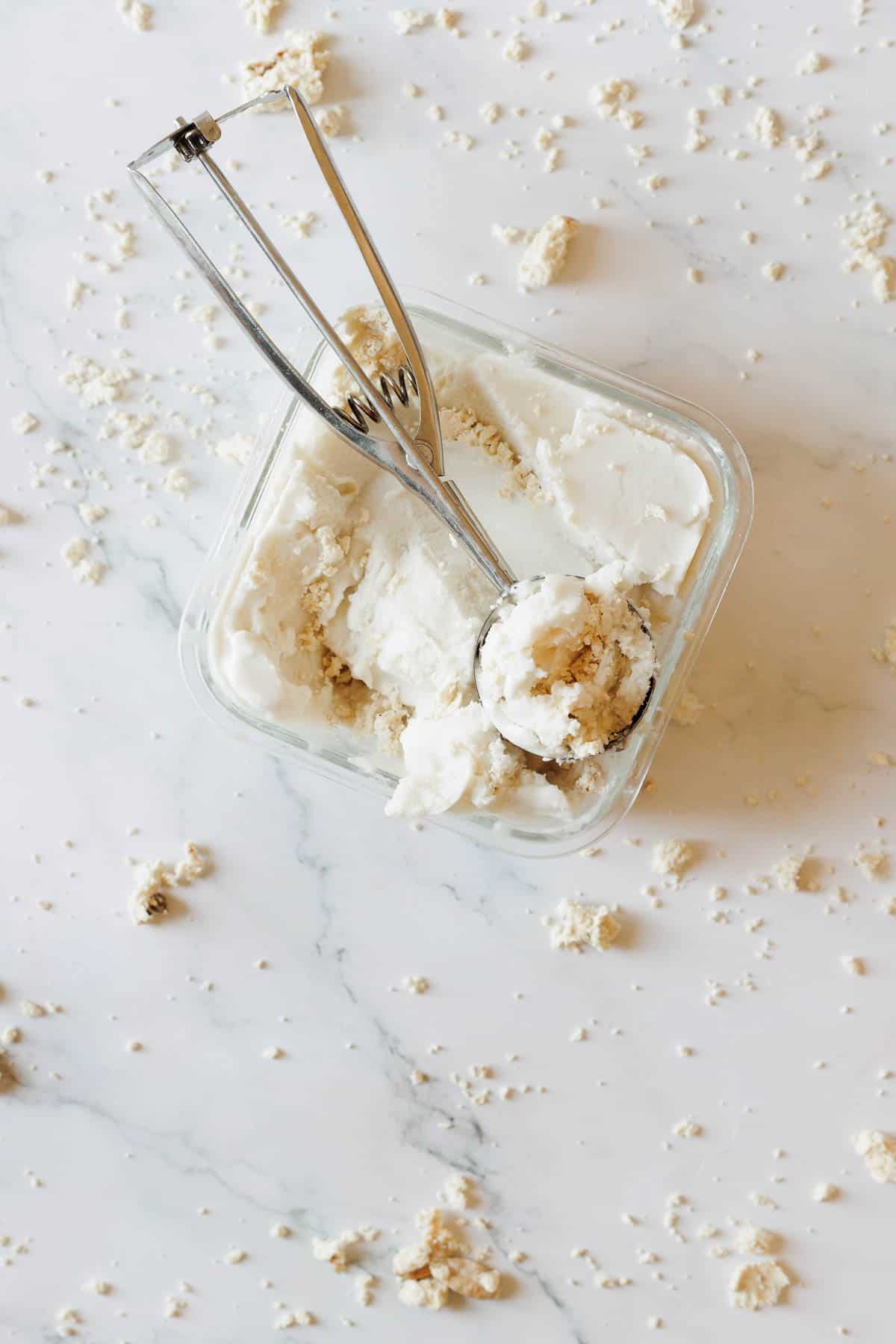 A glass container of Halva Tahini Ice Cream sits on a marble surface, with an ice cream scoop inside. Crumbs are scattered on the ice cream and across the surface.