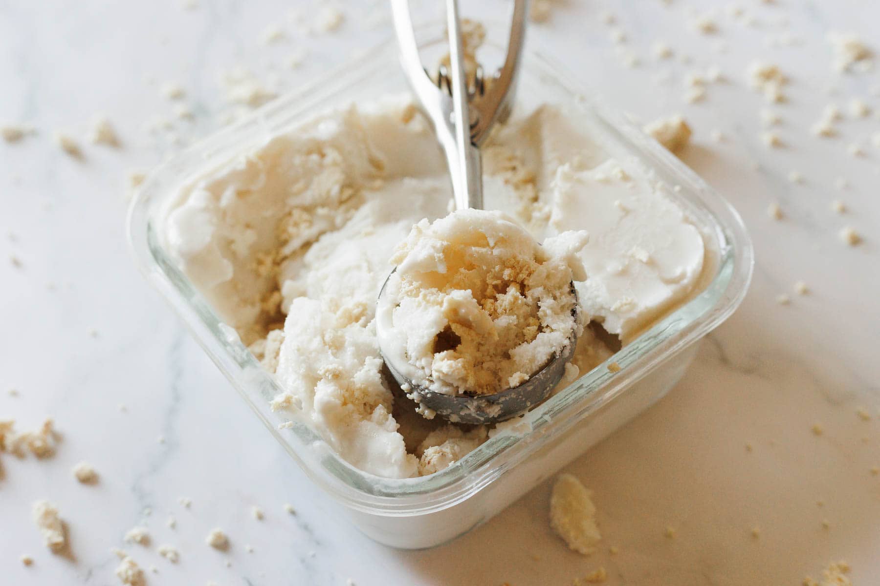 A metal ice cream scoop rests in a glass container filled with white, crumbly Halva Tahini Ice Cream. Crumbs of the unique ice cream flavor are scattered on the white marble surface around the container.