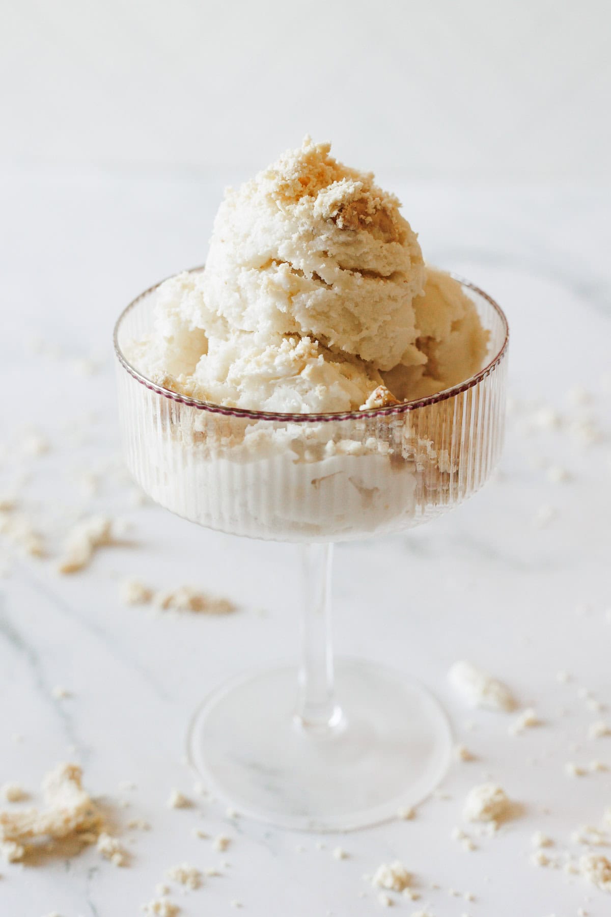 A glass dessert cup filled with scoops of Halva Tahini Ice Cream, topped with light brown crumbs, sits on a white surface with additional crumbs scattered around.