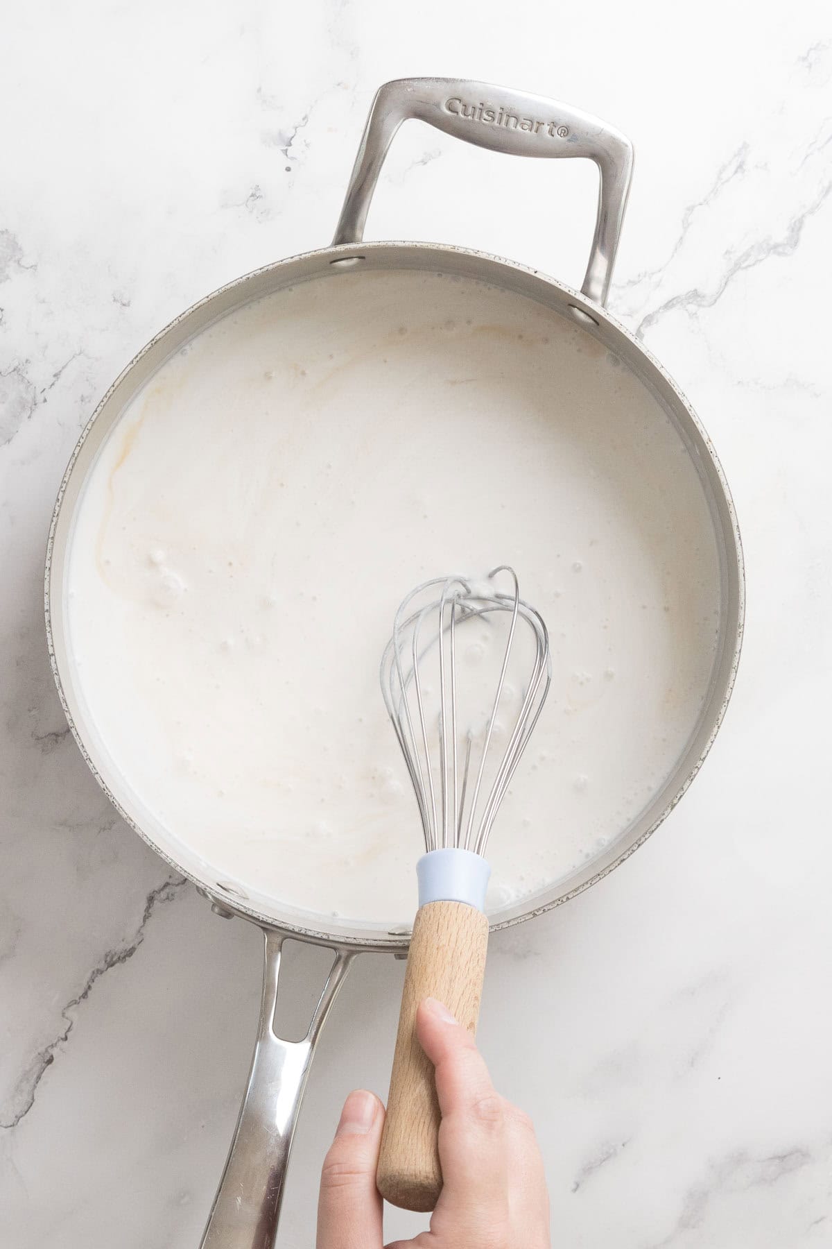 A hand holding a whisk stirs a creamy white Halva Tahini Ice Cream sauce in a stainless steel pan on a white marble surface.