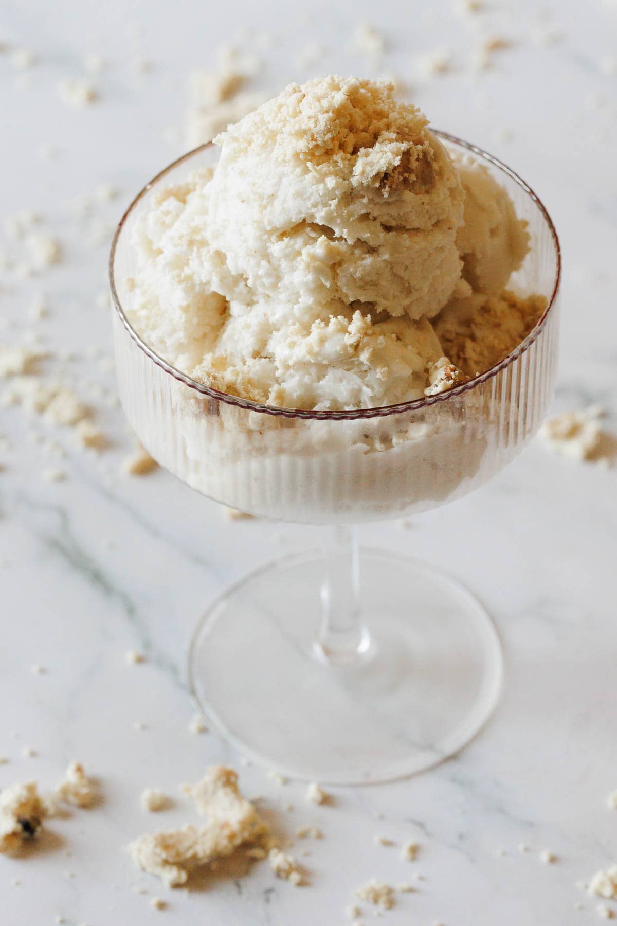 A glass dessert cup filled with scoops of Halva Tahini Ice Cream topped with crumbled cookies, with more crumbs scattered on a white marble surface.