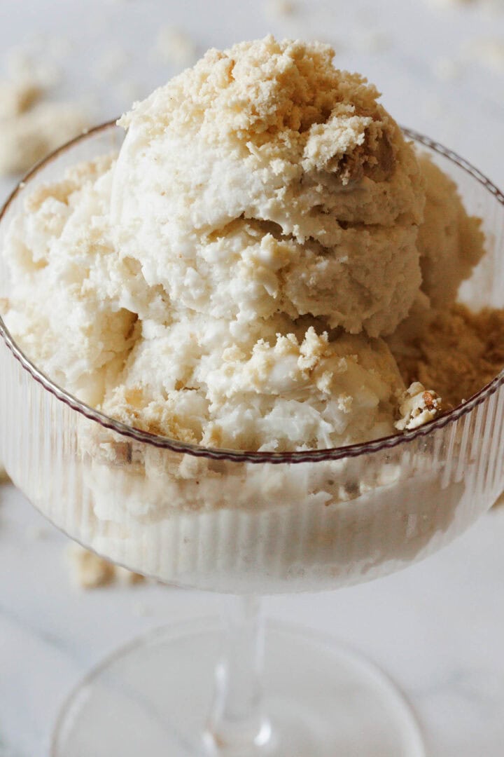 A close-up of scoops of Halva Tahini Ice Cream in a glass dessert dish, topped with crumbled cookie pieces.