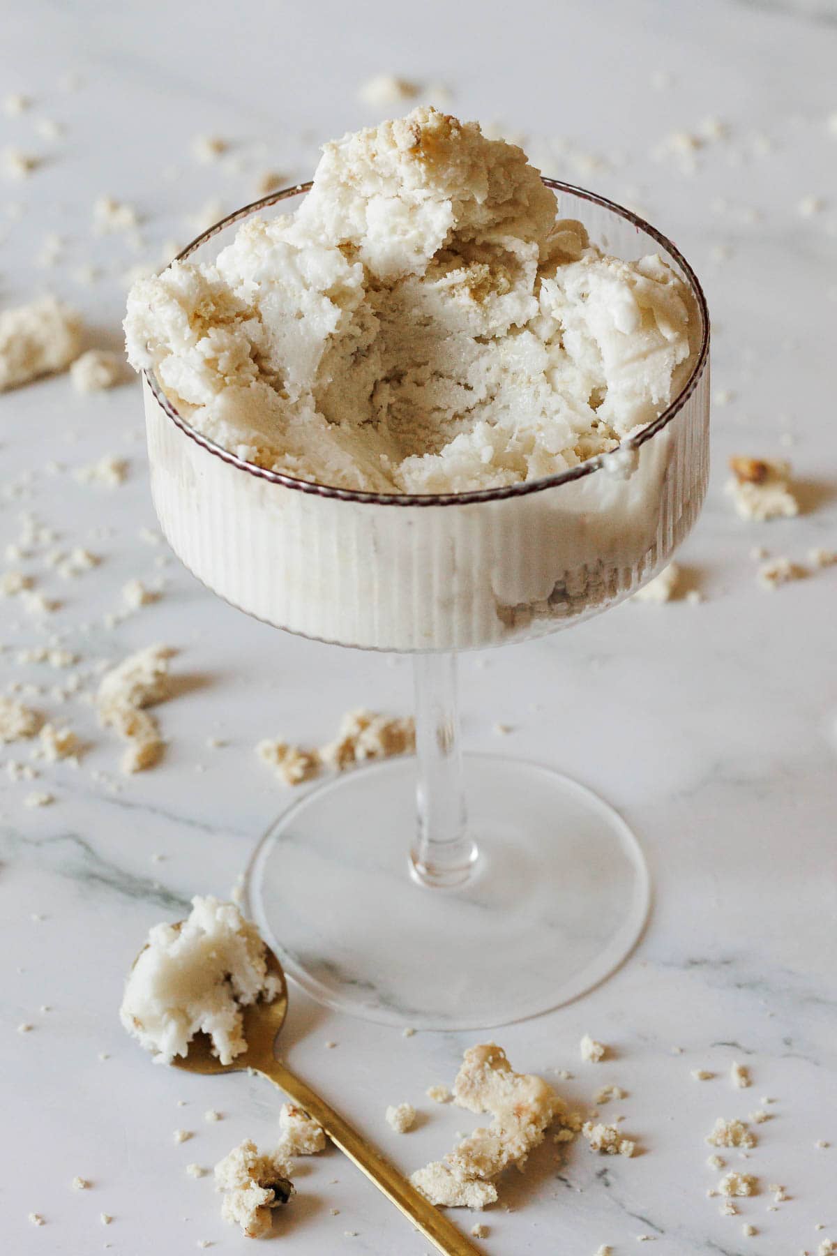 A coupe glass filled with creamy Halva Tahini Ice Cream, topped with crunchy crumbs and surrounded by more on a marble surface. A gold spoon with a small serving of the ice cream rests nearby, ready to enjoy.