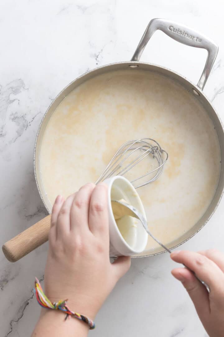 A person holding a small white bowl with a spoon, pouring its contents&mdash;perhaps for Halva Tahini Ice Cream&mdash;into a saucepan of creamy liquid with a whisk inside, on a light marble countertop. The person wears a colorful string bracelet.