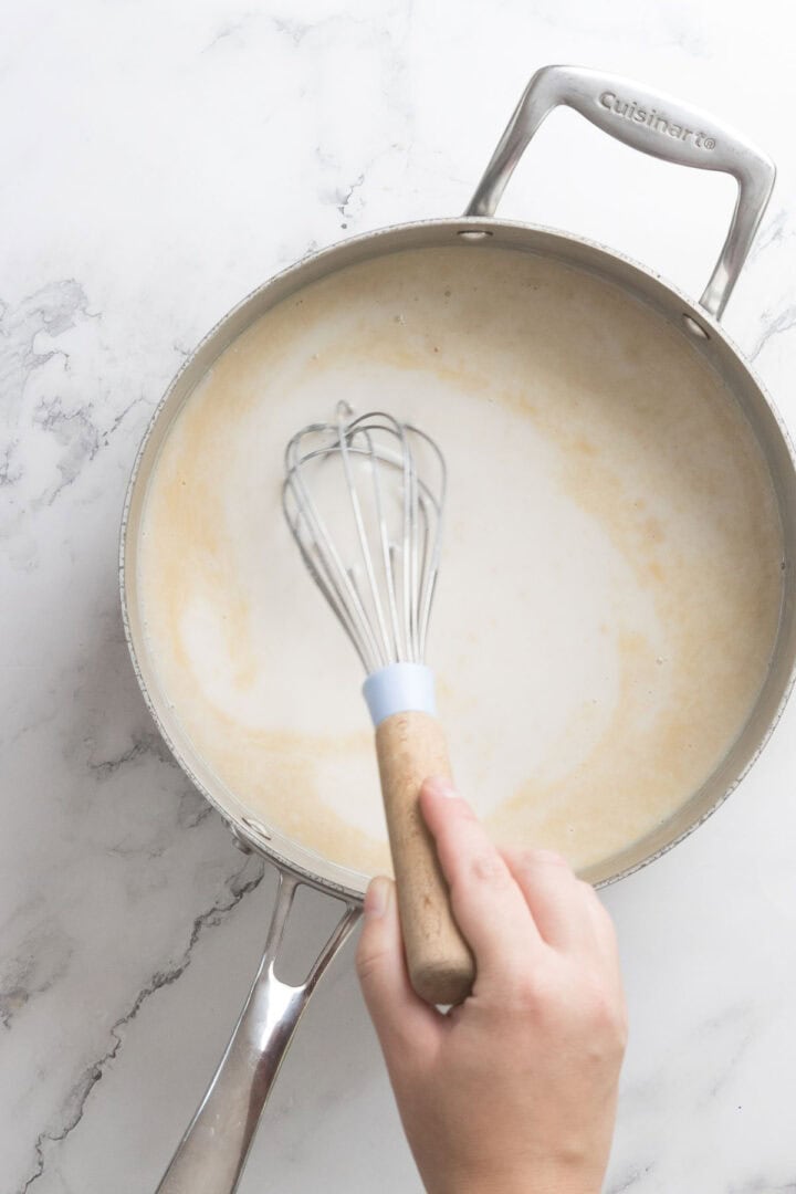 A hand is whisking a light-colored sauce for Halva Tahini Ice Cream in a stainless steel saucepan on a white marble surface.