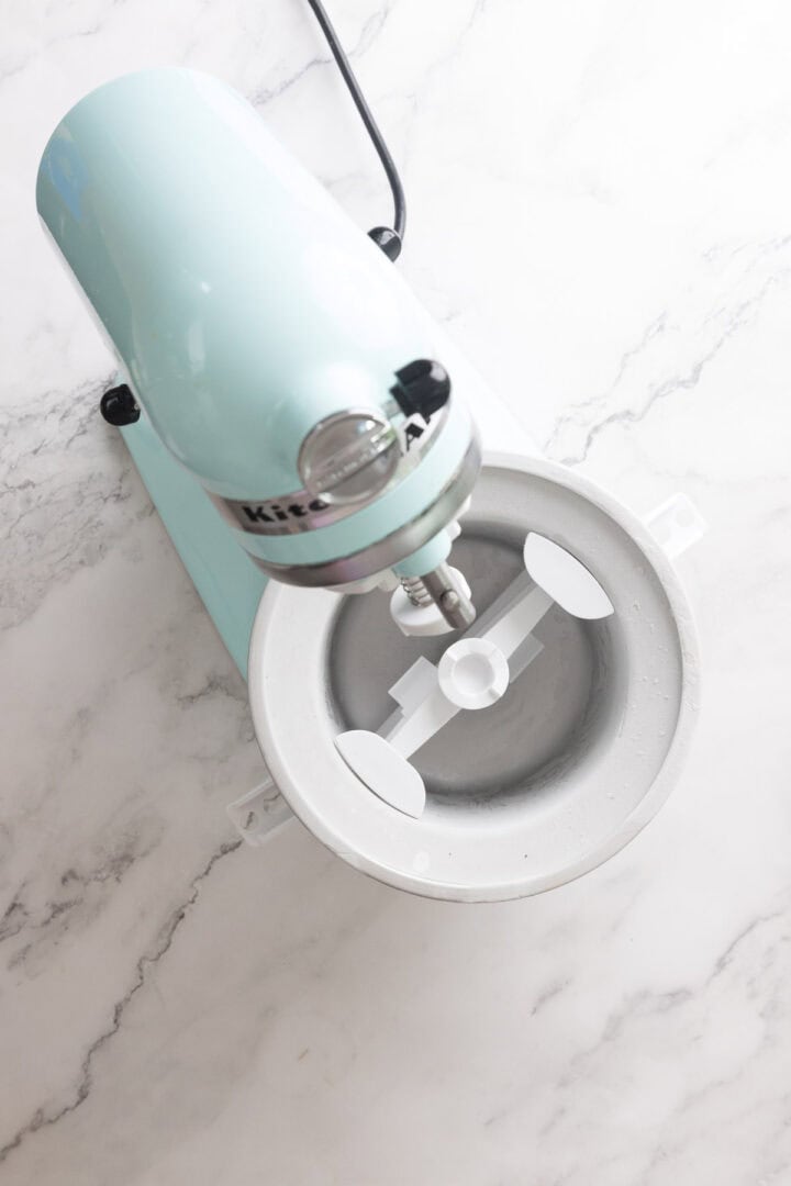 A top-down view of a light blue KitchenAid stand mixer with an ice cream maker attachment on a white marble countertop, ready to churn Halva Tahini Ice Cream.