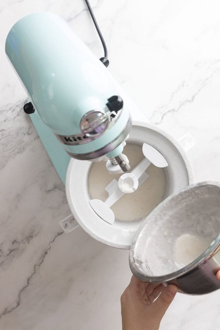 A person pours a Halva Tahini Ice Cream mixture from a metal bowl into a stand mixer with a white attachment on a marble countertop.