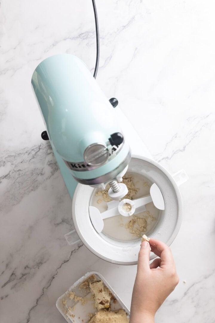 A hand drops a piece of dough or mixture for Halva Tahini Ice Cream into the bowl of a light blue stand mixer on a white marble countertop. Mixing attachments are visible inside, with a container of dough nearby.