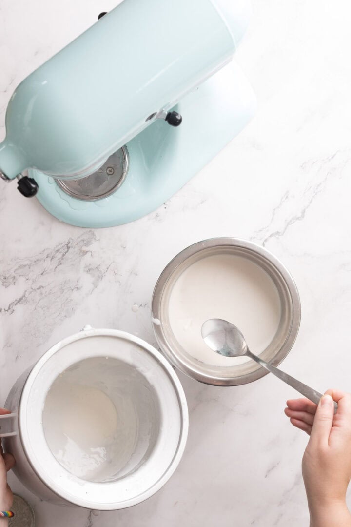 A person holds a spoon over a metal bowl of white liquid&mdash;perhaps Halva Tahini Ice Cream&mdash;next to another bowl and a light blue stand mixer on a marble countertop.