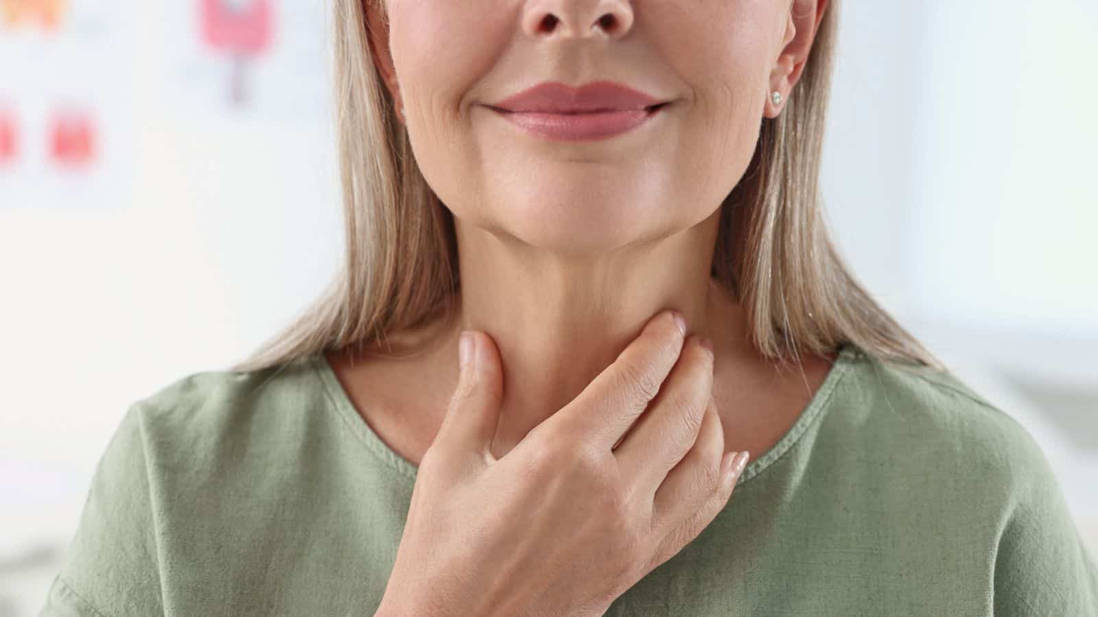 A woman wearing a green shirt gently touches her throat with her hand. She is smiling softly, and the background is softly blurred, focusing attention on her face and hand.