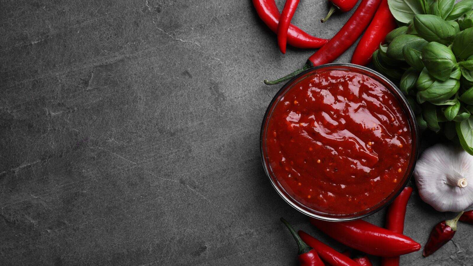 A bowl of red chili sauce sits on a dark stone surface, surrounded by fresh red chili peppers, basil leaves, and a head of garlic. The ingredients are arranged closely beside the bowl on the right side of the image.