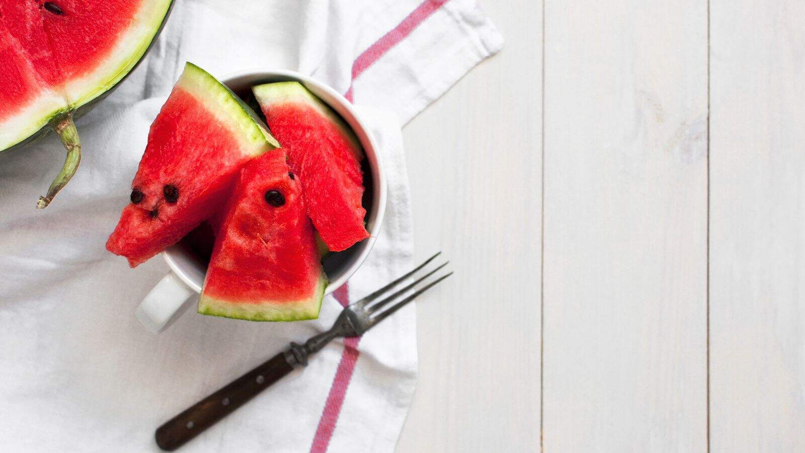 A white cup filled with watermelon slices sits on a white cloth with a pink stripe. A fork lies nearby, and part of a watermelon is visible in the upper left corner on a light wooden surface.